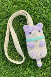 The finished purple and beige crochet cat phone bag with its long strap lying flat on green grass